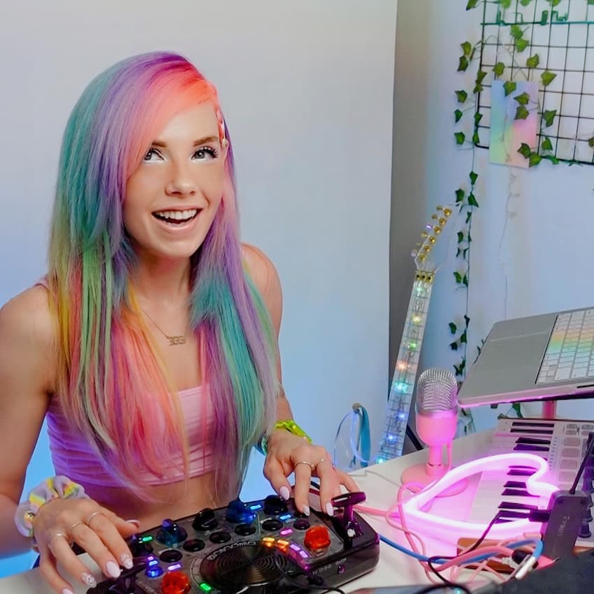 Musician with rainbow hair smiling while playing a Blipblox After Dark synthesizer in a colorful home studio.